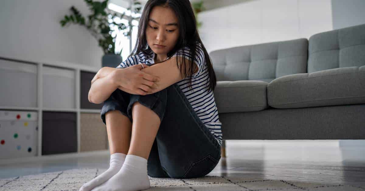 Teen girl sitting on the floor, hugging her knees, looking depressed.