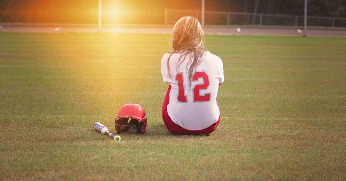 Upset teen girl sitting on a field with a softball helmet and bat beside her