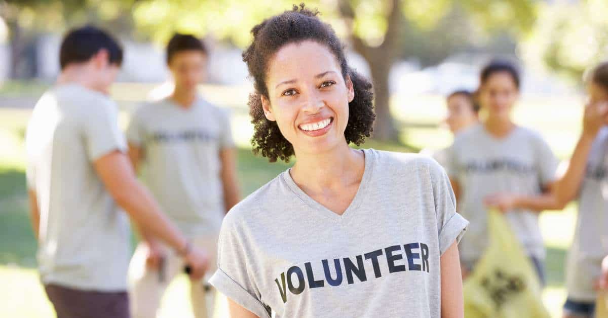 Teen with a shirt that reads, "volunteer" stands in the foreground. Behind her, other teens are filling garbage bags with litter.