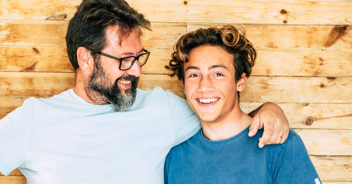 Father looking at his son with his arm around him while son looks at the camera.