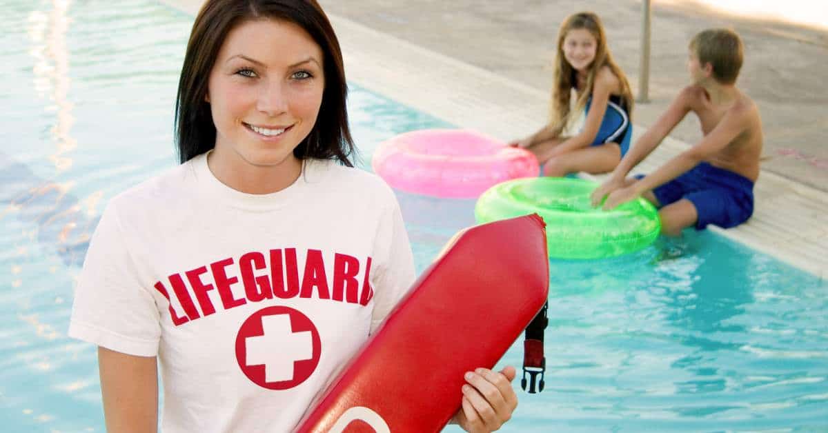 Female teen lifeguard in the foreground with two younger kids in a pool in the background.