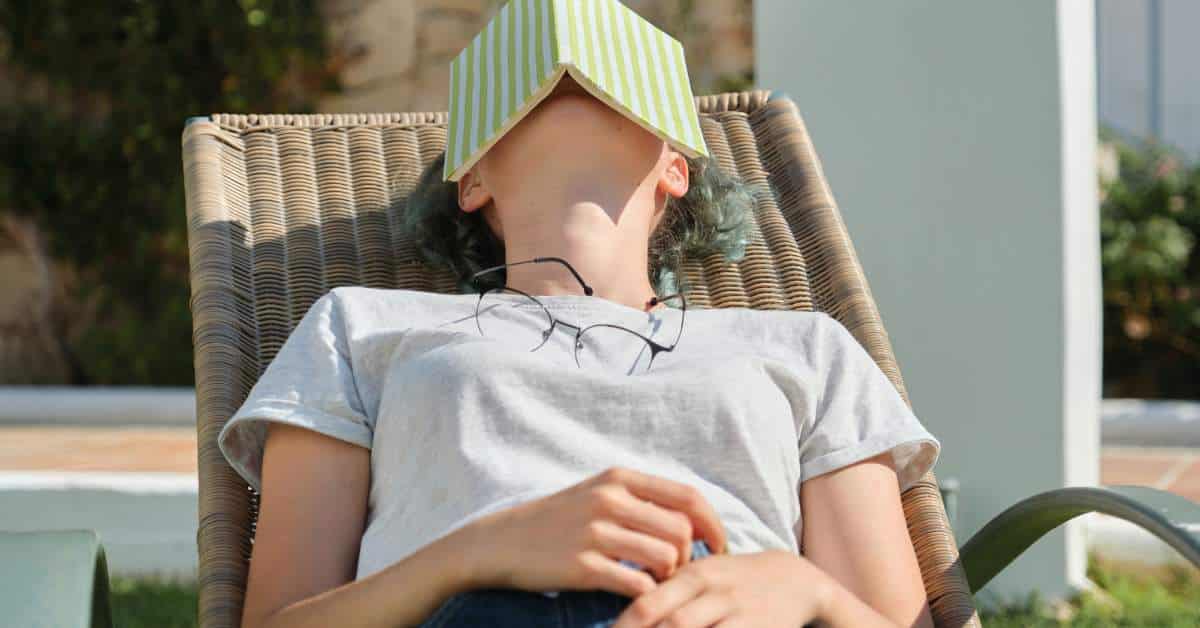 Teen lying on a lawn chair, reclined, with a book over her face and her glasses off.