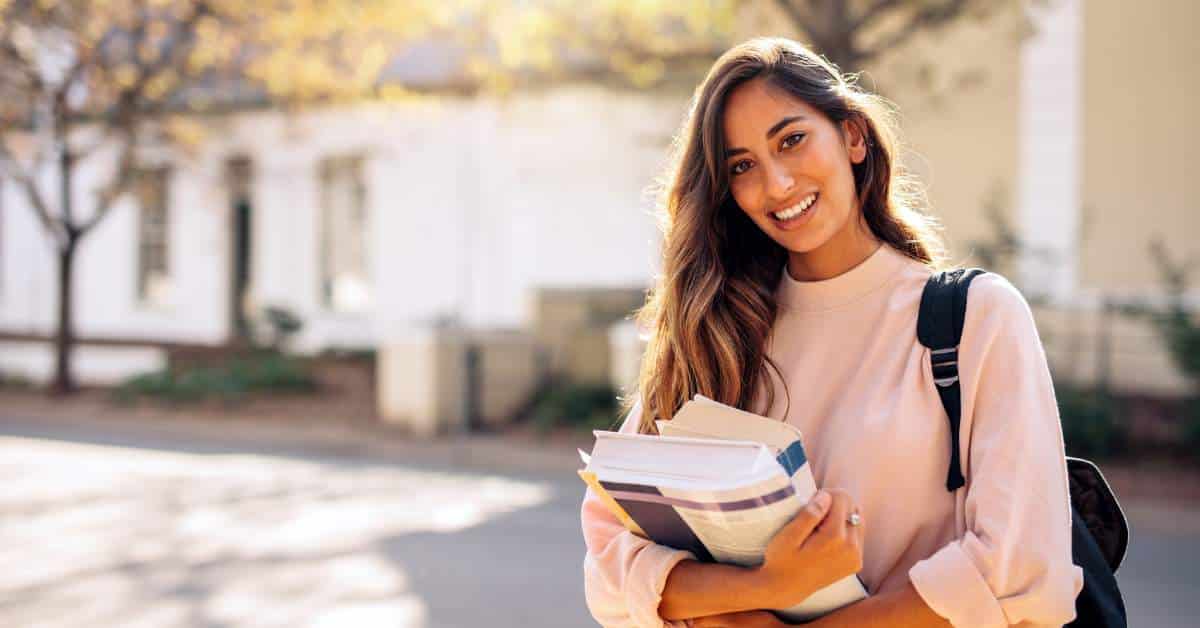 Young woman with long hair holding books in her arms standing on a college campus.