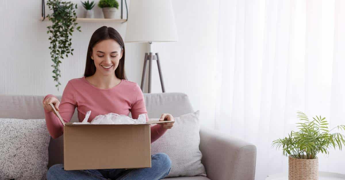 Young woman sitting on a couch opening a box. Her face shows surprise and happiness.