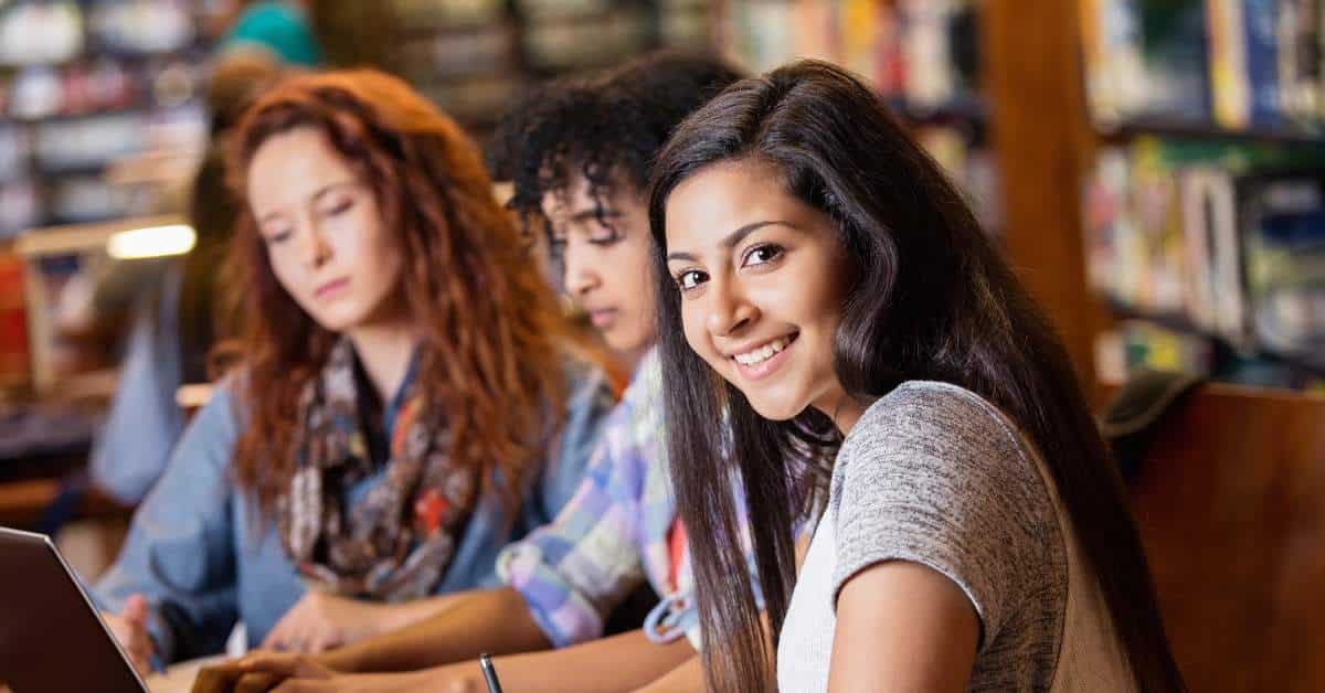 Three teen girls studying together in the library with one teen girl smiling at the camera