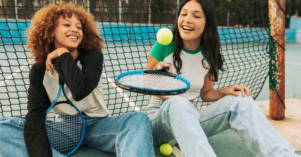 Two teen girls sitting by a tennis net holding tennis rackets