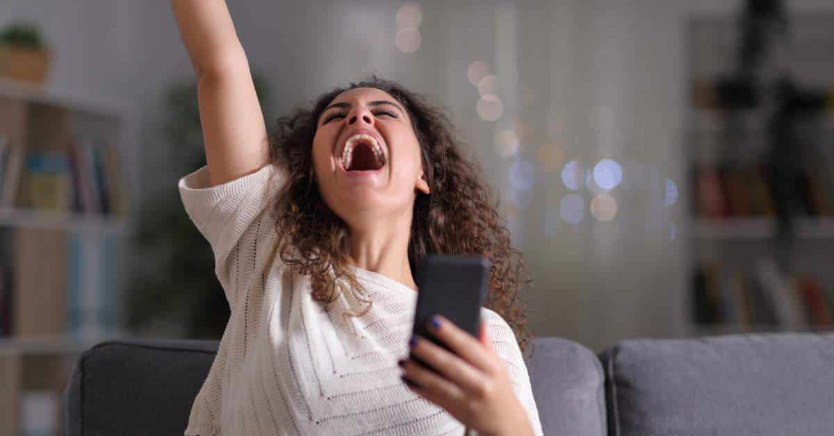 Young woman holding her phone in one hand, raising her other arm in victory. Her head is back, and she is excitedly exclaiming.