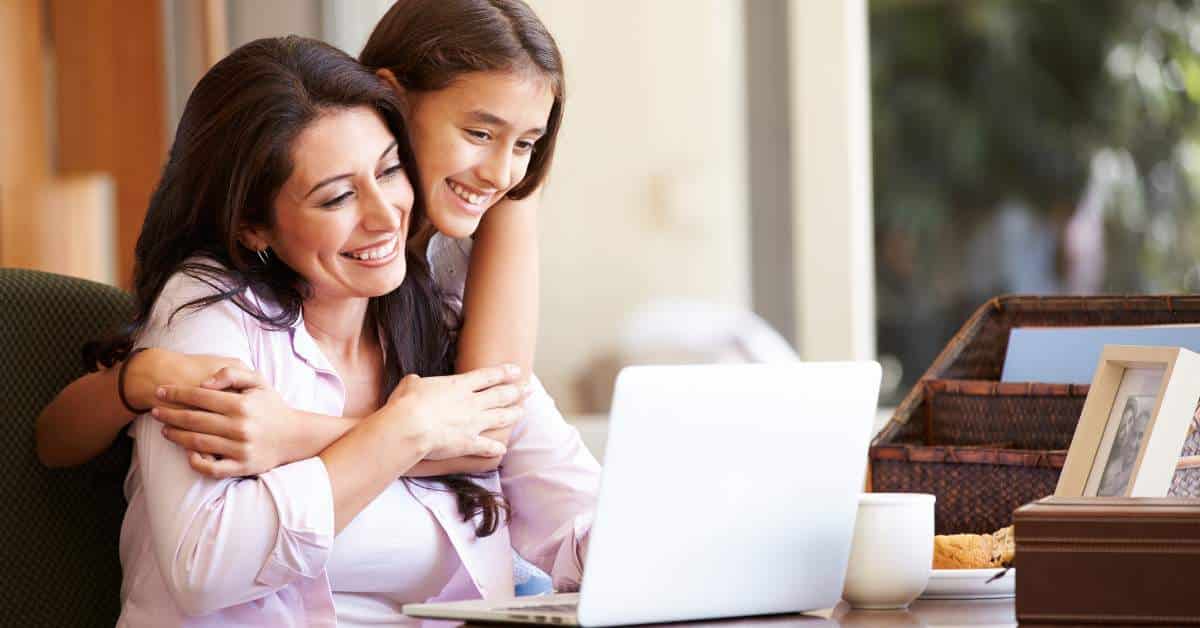 Mother sitting in a chair in front of an open laptop on a desk. Her teen daughter is at her side hugging her.