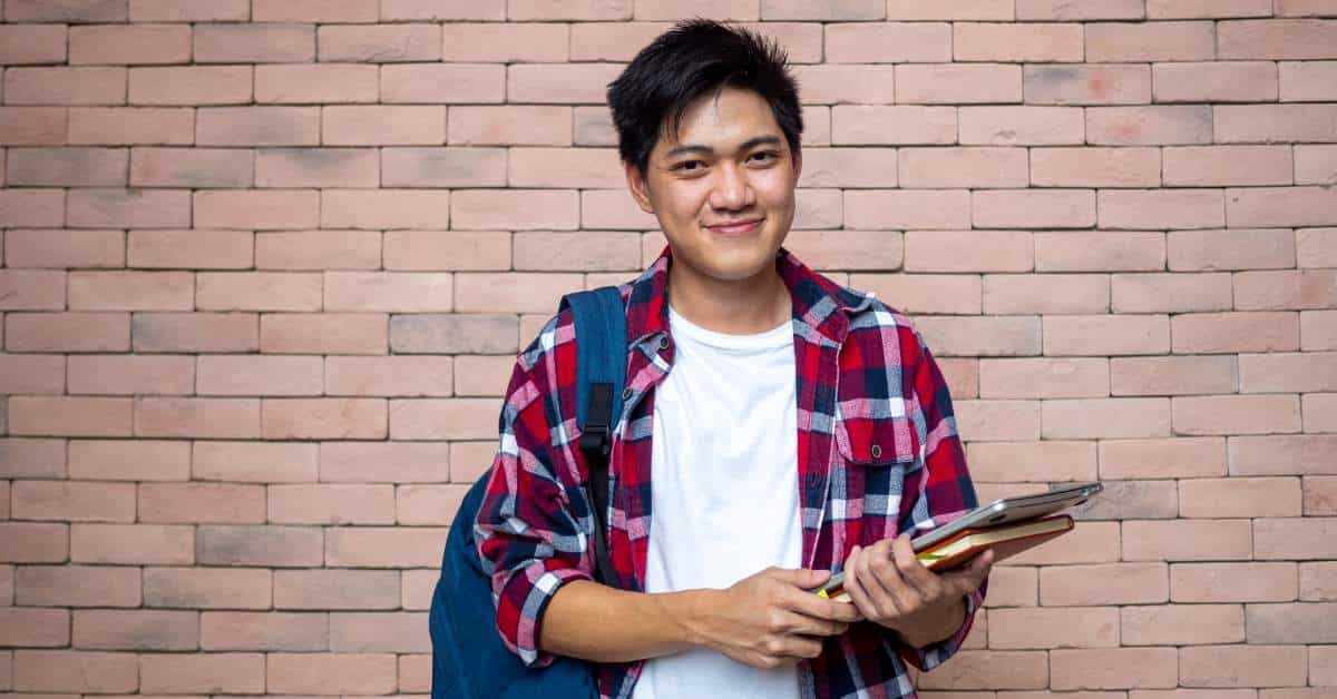 Smiling young man holding a notepad and standing against a brick wall.