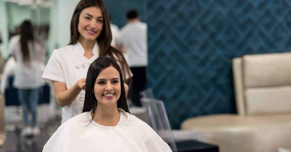 A young woman hairdresser styling a young woman's hair. Both are smiling at the camera