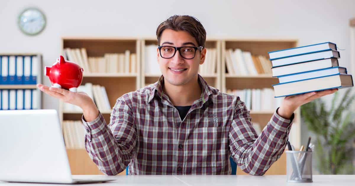 Young man with glasses sitting at a table holding books in one hand and a piggy bank in the other.