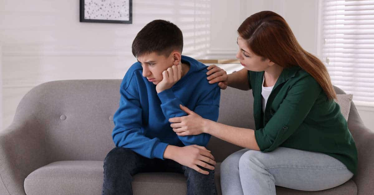 Dejected teen son sits with his face away from his mother while she tries to talk to him and comfort him.
