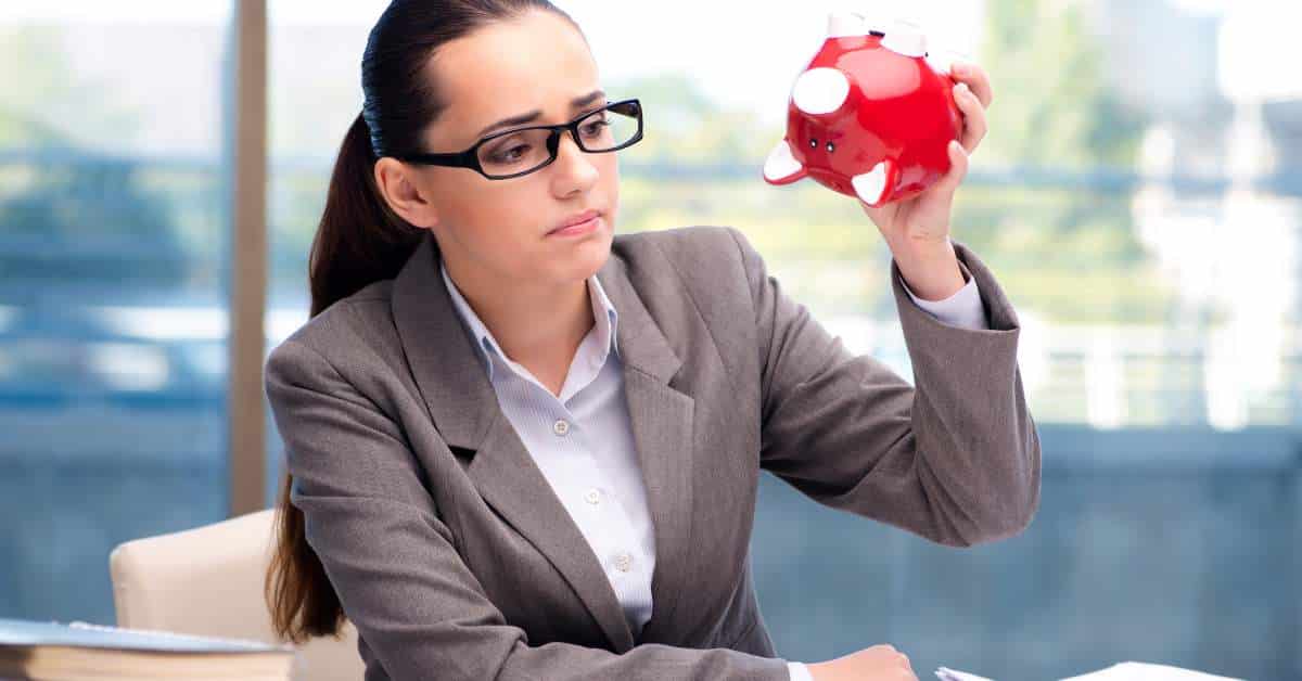 Woman dressed in a suit wearing glasses sitting at a desk turning a red piggy bank upside down.